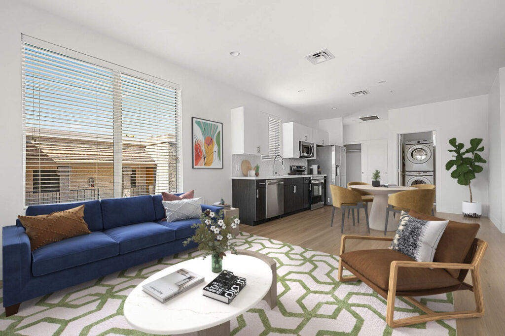 living room with large 2 panel wall windows, a blue couch and white center circle table, view of the kitchen and stacable washer and dryer.