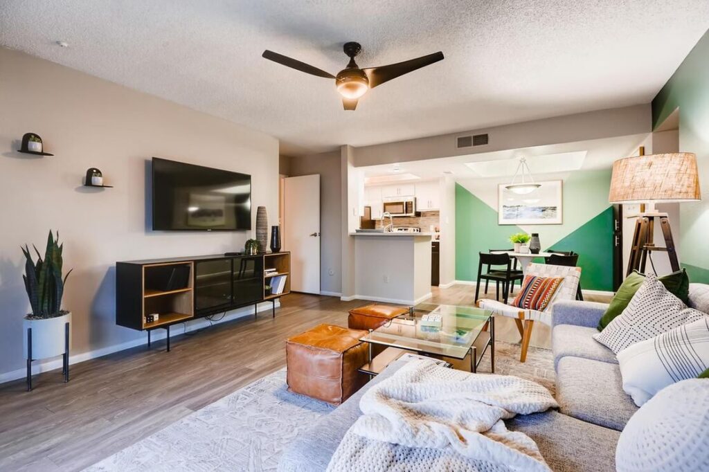 view of living room with slight view of dining and kitchen area. TV mounted on the wall with 3 arm ceiling fan and a decorative plant. rectangular glass center table