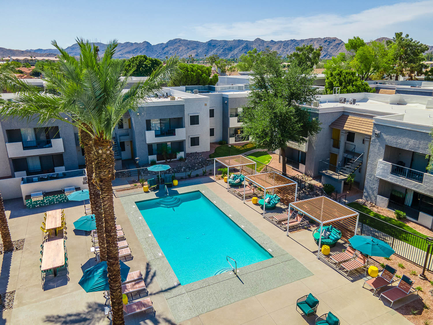 drone shot of swimming pool and the overlooking buildings that surround the pool. 3 semi covered cabanas, lounge chairs and seating areas